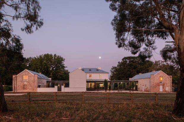 Twilight exterior view of a symmetrical contemporary residential property, comprising a central pale rendered two-storey house flanked by two low brick pavilion-like wings. The buildings have simple pitched roofs, dark-framed glazing, and warm interior lighting. Mature eucalyptus trees, a timber fence, and a rural-looking foreground landscape frame the composition, with a visible moon in the dusky sky.