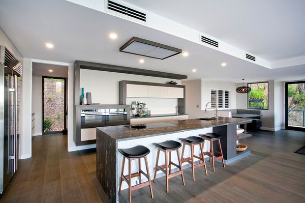 Contemporary open-plan kitchen with a large dark stone island, four bar stools, sleek white and dark cabinetry, integrated appliances, timber-look flooring, recessed downlights, and a linear ceiling feature. Large windows and glazed doors provide abundant natural light and views of greenery.