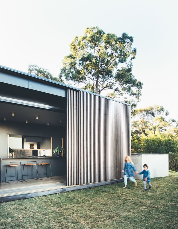 Exterior view of a contemporary single-storey pavilion-like home with a dark open entertaining area, bar-height outdoor counter and stools, and a prominent vertical timber batten screen. The building opens onto a lawn and landscaped suburban setting, with children visible in the foreground.