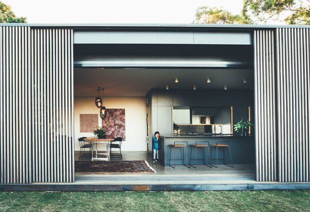 Front-on view of a low, modern residential building with a wide open glazed frontage and vertically ribbed dark cladding. The interior visibly combines a dining area on the left with a compact kitchen and breakfast bar on the right. Strong horizontal roofline, large opening, dark cabinetry, light walls, and vertical facade fins create a clean contemporary composition.
