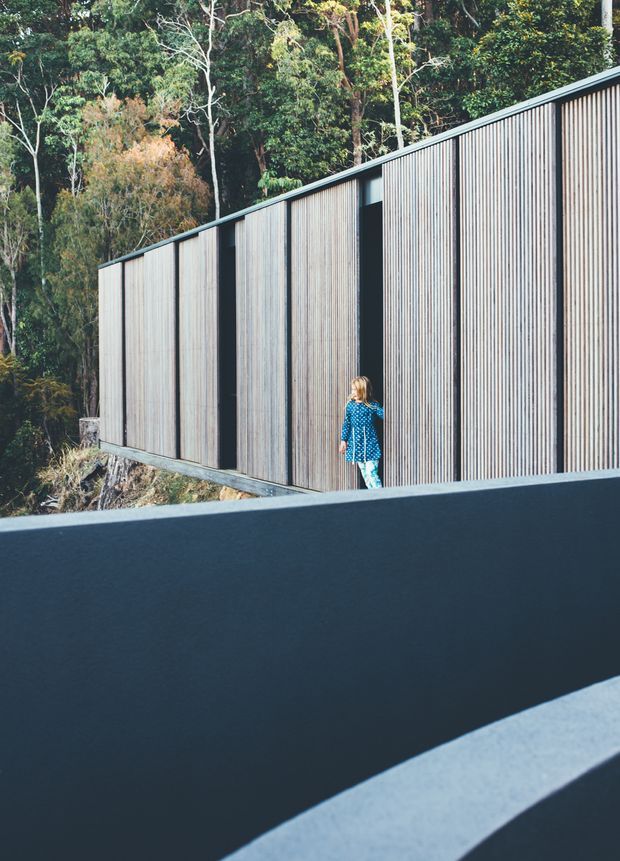 Exterior architectural photograph of a long, narrow modern building set into a densely wooded, sloping site. The facade is composed of closely spaced vertical timber-toned battens or cladding, with dark recessed openings and a simple sloping roofline. A dark curved or angled foreground structure partially obscures the lower building, while a person provides scale.