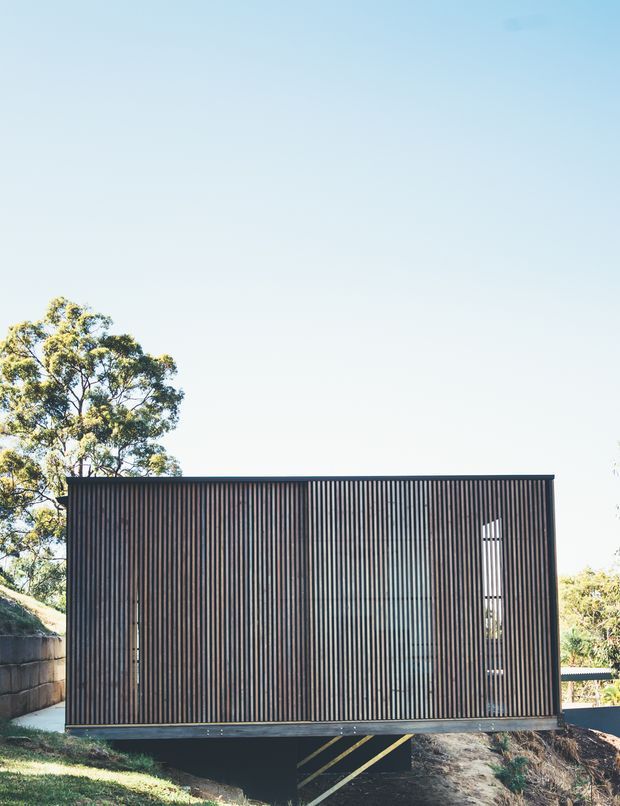 A compact contemporary dark-clad building viewed from outside, set on a sloping site among mature trees. The facade is dominated by closely spaced vertical timber-like battens, with narrow vertical glazed openings and a raised lower section beneath the main volume. Clear blue sky and landscaped surroundings frame the minimalist exterior.