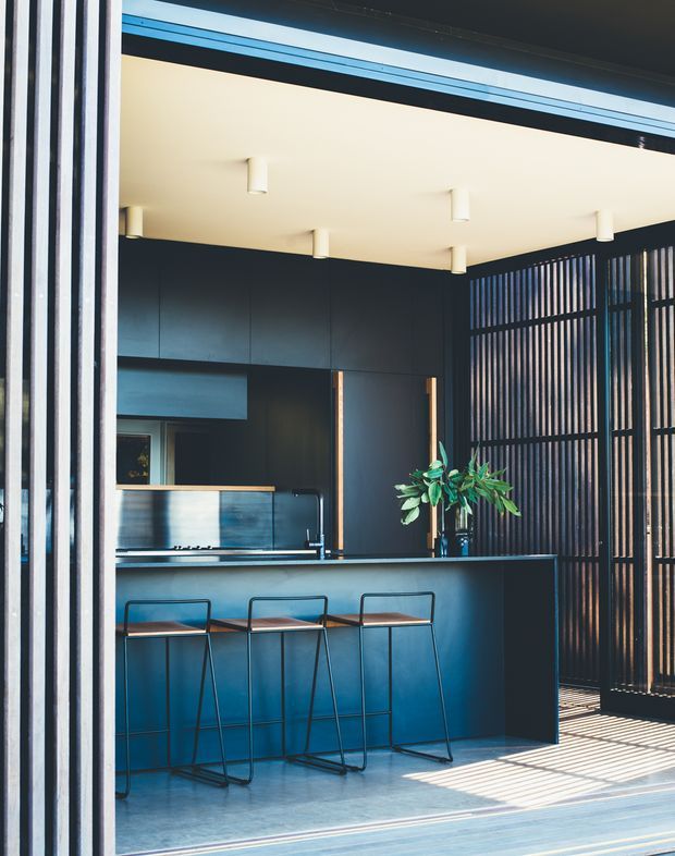 A sleek, dark-toned contemporary kitchen viewed from an open glazed doorway. The room features a large island with three bar stools, dark blue-black flat-panel cabinetry, a matching benchtop, integrated appliances, and a reflective metallic splashback. A vertical timber-like slatted screen forms the right side of the space, casting strong linear shadows across the pale floor. The ceiling is light with simple cylindrical downlights, creating a high-contrast, refined interior.
