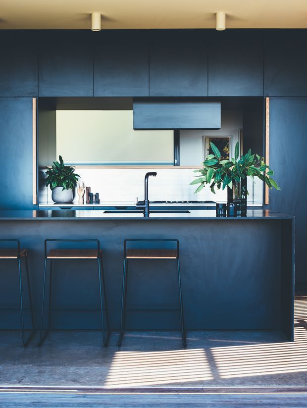 A compact, sophisticated contemporary kitchen dominated by deep blue cabinetry and a matching island. The space features a dark benchtop, integrated rangehood, black tapware and appliances, timber-accented bar stools, indoor plants, and strong linear sunlight filtered through blinds across the floor and island.