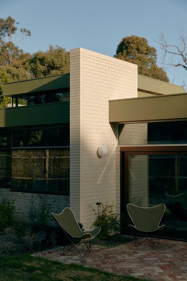 Exterior view of a modern residential building with a pale cream brick feature wall, dark green flat-roofed volumes, extensive dark glazing, and timber-framed doors. A brick-paved patio with two sculptural lounge chairs sits in the foreground, surrounded by established trees and planting.