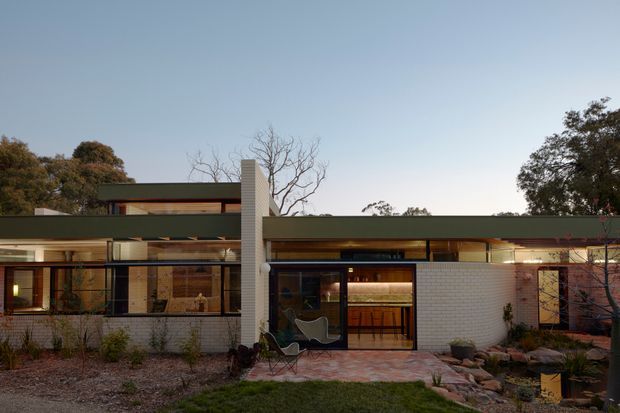 Single-storey modern residence viewed from the landscaped front garden at dusk. The low horizontal building has broad glazing, dark window frames, pale brick walls, a projecting flat roof, and a prominent narrow vertical brick element. Warm interior lighting is visible through the windows and entry glazing, while the foreground includes a paved path, gravel garden beds, and established trees.