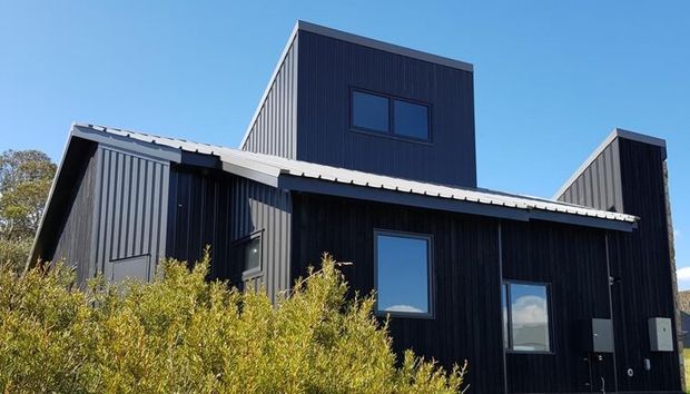 Exterior view of a contemporary dark-clad building with multiple intersecting pitched roof volumes, vertical ribbed facade panels, and simple rectangular windows and doors. The structure is set among dense shrubs under a clear blue sky.