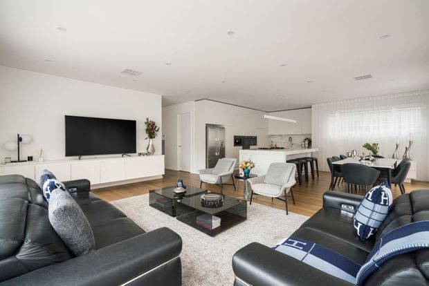 Bright, open-plan contemporary living and dining area with white walls and ceiling, pale timber flooring, and large sheer-covered windows. Two black leather sofas frame a glass coffee table and neutral rug, while a white entertainment unit with wall-mounted television sits to the left. The rear kitchen includes a compact island or peninsula with stools, and the dining area has a dark table and chairs. Minimal recessed lighting and streamlined cabinetry create a clean, spacious interior.