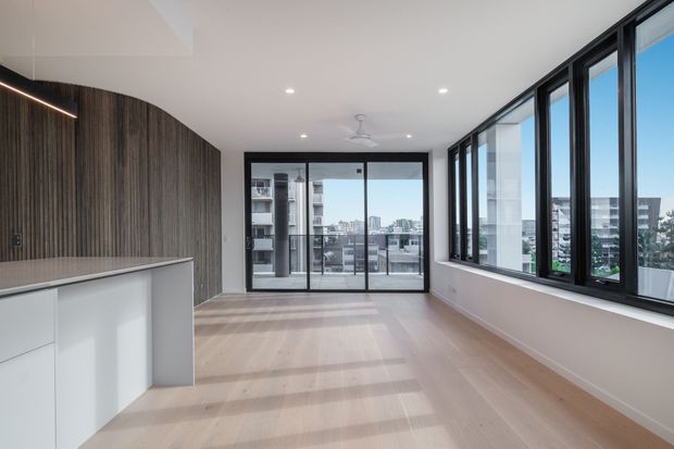 Bright, contemporary open-plan apartment interior with pale timber flooring, a white kitchen island, dark vertically grained timber wall panelling, and expansive black-framed glazing overlooking an urban setting. The room is largely unfurnished and could function as a living or dining area.