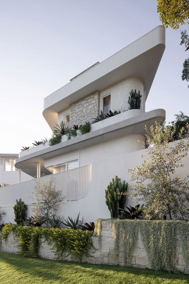 Photograph of a refined two-storey residential exterior with sweeping curved balcony edges, a broad rounded roof canopy, pale rendered walls, vertical ribbed cladding and a textured light stone feature wall. Layered balconies are planted with cacti and drought-tolerant greenery, while a low stone garden wall and manicured lawn frame the facade. The design has a sculptural, coastal-modern character with strong horizontal lines and rounded corners.