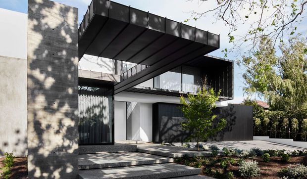 Exterior view of a contemporary multi-level residence with strong cantilevered forms, dark metal cladding, extensive glazing, a projecting upper terrace or roof plane, and a tall textured concrete or masonry wall. The entry is approached by broad steps through a landscaped front garden, with mature trees casting shadows across the facade.