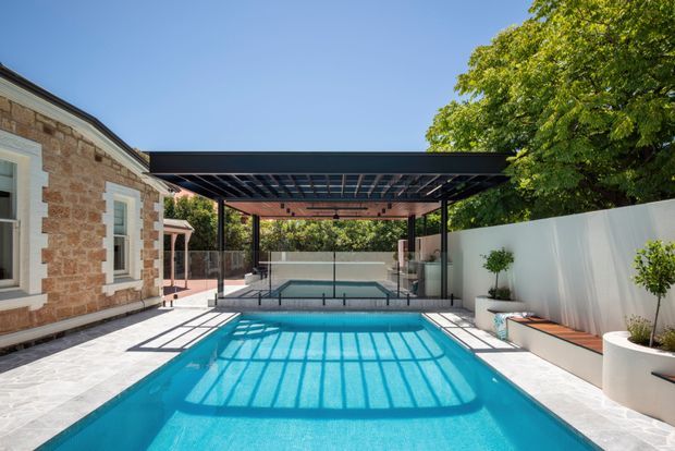 Bright outdoor residential pool area with a long rectangular swimming pool in the foreground, covered by a black steel pergola with a translucent or open slatted roof structure. A smaller raised spa or secondary pool sits beyond a glass balustrade. The area is paved in pale stone, bordered by a white rendered boundary wall with built-in bench seating and planters. A brick masonry house with white-trimmed windows is visible on the left, with established trees beyond.