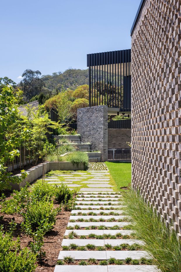 A landscaped residential exterior walkway with large pale stepping slabs set through lawn and planted garden beds. A tall textured brick wall dominates the right side, while contemporary dark metal vertical screening and brick structures frame the path. The garden slopes gently toward a wooded suburban backdrop under clear daylight.