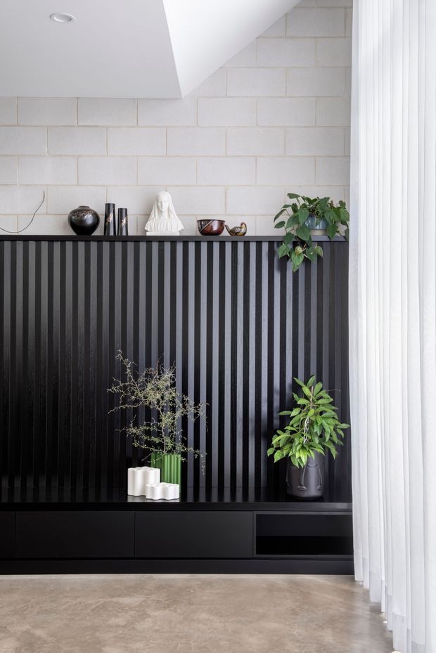 A refined interior feature wall with a black vertical-ribbed built-in joinery unit beneath a pale rendered masonry wall. Decorative objects and potted plants sit on the shelving, while sheer white curtains frame the right side. The space has a polished, contemporary feel with a neutral palette and strong black-and-white contrast.