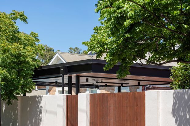 Exterior view of a contemporary covered outdoor entertaining area behind a white rendered boundary wall and warm timber gate. The structure has a broad flat roof with dark metal fascia and posts, timber-lined soffits, and recessed or track-style lighting. Mature leafy trees partially screen the building, with a pitched-roof house visible behind.