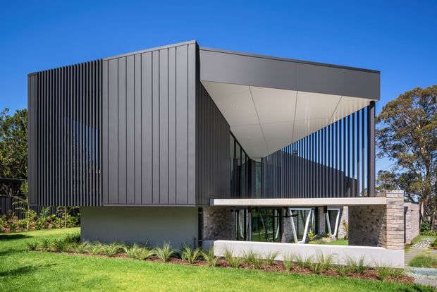 Contemporary two-storey residence set in a landscaped suburban setting, featuring a strongly geometric cantilevered upper volume, dark vertical fins, grey metal cladding, extensive glazing, and a light stone-clad lower level. A dramatic angled soffit creates a sheltered terrace or entry zone beneath the upper floor.