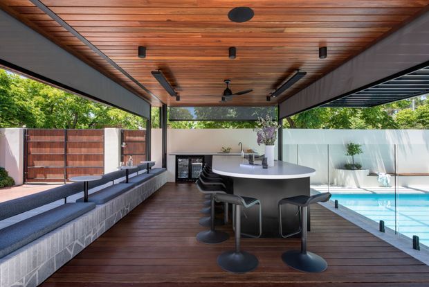 A covered outdoor entertaining pavilion beside a swimming pool, featuring a dark timber-lined ceiling with exposed linear boards, integrated strip lighting and ceiling fans. The space includes a curved island bar with stools, built-in bench seating along a tiled masonry wall, a compact outdoor kitchenette, timber decking, and large open screened sides overlooking the pool and garden.