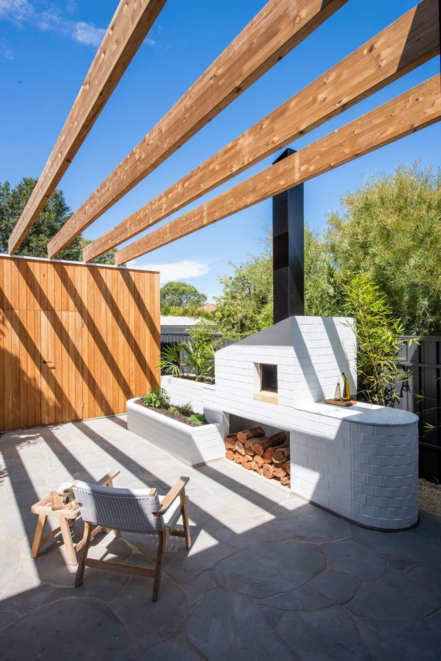 Sunny landscaped courtyard patio with a built-in white-painted brick outdoor fireplace and adjoining curved bench or counter. Timber privacy fencing, exposed timber pergola beams, dark structural post, paved flooring, raised planter beds, and outdoor lounge chairs create a warm contemporary outdoor entertaining space.
