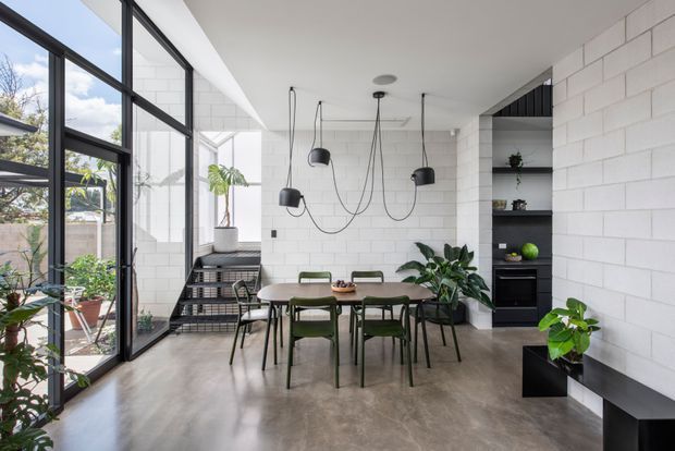 A bright contemporary dining area with a concrete-look floor, white painted block walls, full-height black-framed glazing, and a sculptural cluster of black pendant lights. A dark built-in shelving and appliance wall sits behind the dining table, while indoor plants and a small courtyard are visible through the glazing. Green dining chairs and a timber-toned table add restrained colour to the monochromatic interior.