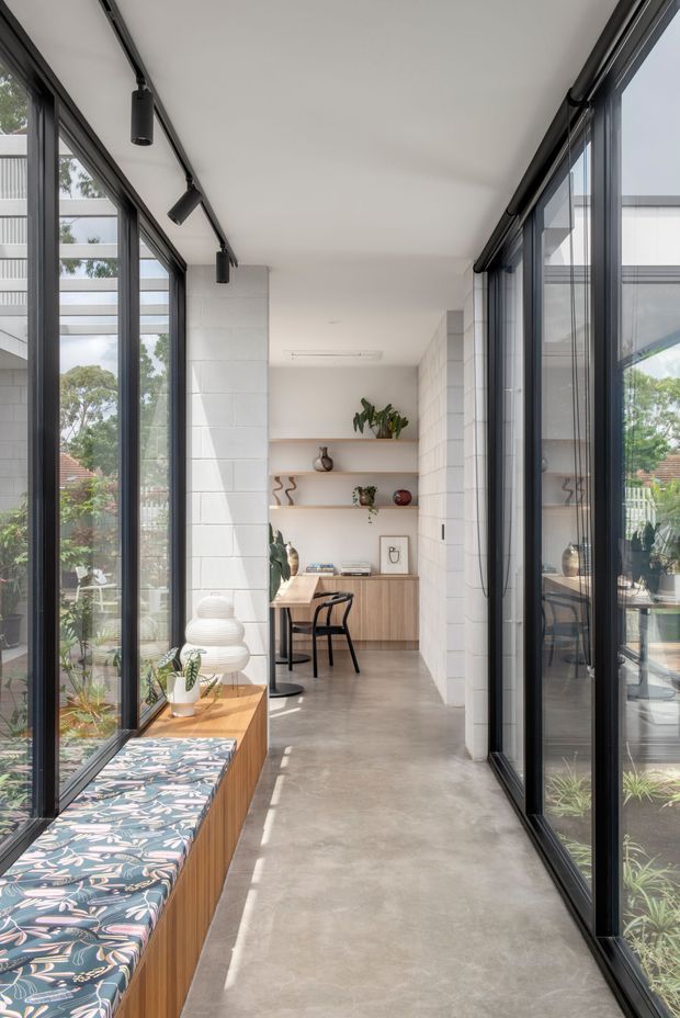 A bright, narrow glazed interior passage with full-height black-framed windows on both sides, a polished concrete floor, white painted block walls, and a built-in timber window seat with patterned upholstery. The corridor leads to a compact dining or work area with a timber table, black chairs, open shelving, cabinetry, plants, and decorative objects. Black track lighting runs along the white ceiling, and garden views are visible through the glazing.