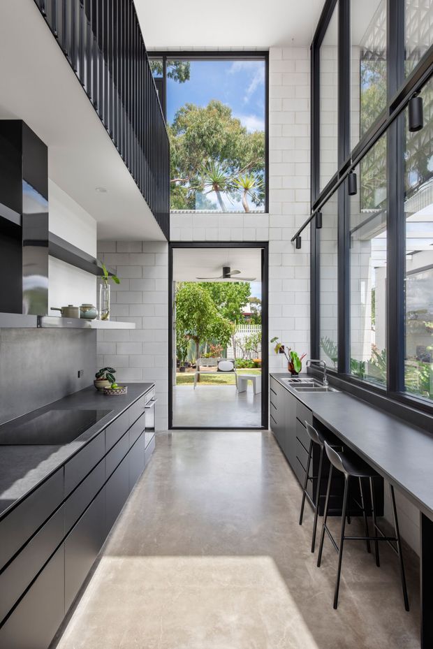 A tall, narrow contemporary kitchen with a dramatic double-height volume. Matte black cabinetry and dark benchtops line both sides, with a long breakfast counter and bar stools beneath expansive black-framed glazing. White rectangular wall tiles, polished concrete flooring, a black vertical balustrade, and clerestory-style windows create a crisp, light-filled interior with strong views to a leafy courtyard.