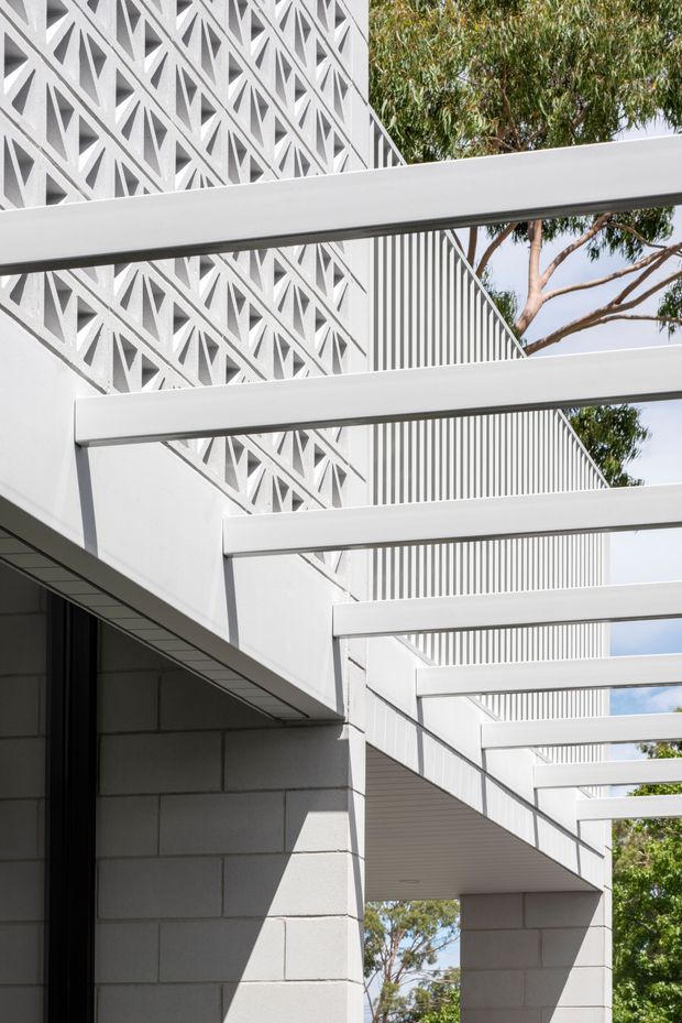 Architectural exterior detail of a bright white modern building, featuring repeated horizontal pergola beams, a patterned breeze-block screen, vertical privacy slats, and pale masonry block walls. Trees and sky are visible beyond the open structure.