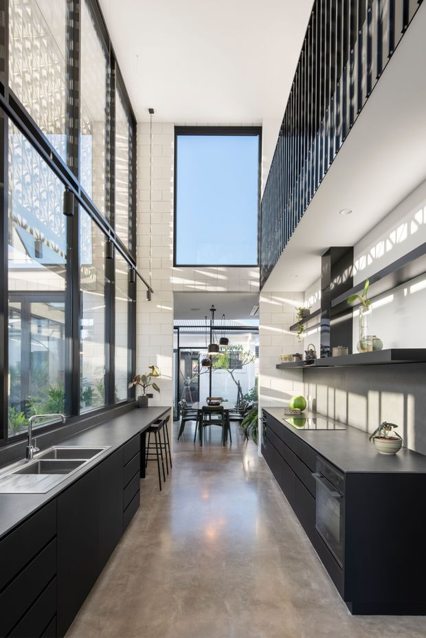 A tall, narrow contemporary kitchen with a dramatic double-height volume, polished concrete floor, extensive black-framed glazing, and a large clerestory-like window. Matte black cabinetry and dark benchtops run along both sides, with stainless-steel sinks, open shelving, integrated appliances, and a compact dining area beyond. A black metal balustrade overlooks the kitchen from an upper level, while strong daylight creates patterned shadows across the white tiled walls.