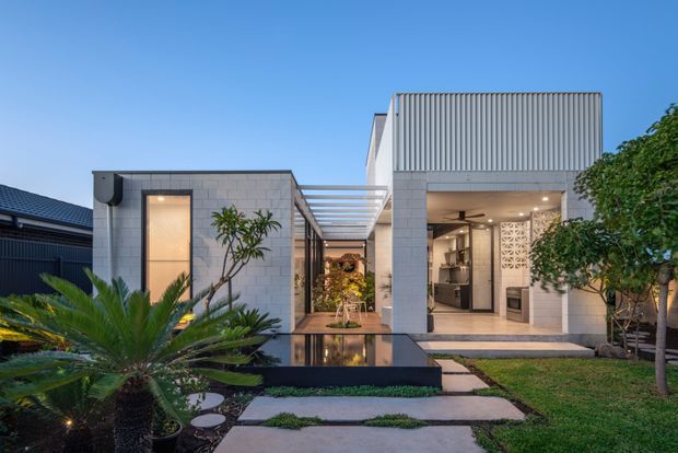 A contemporary single-storey home viewed from the landscaped front garden at dusk. The facade combines pale masonry block walls with a tall ribbed metal-clad volume, broad glazed openings, and a covered linear walkway/pergola leading toward a warmly lit interior. Stepping-stone paths, a shallow reflecting pool or dark water feature, palms, and manicured planting frame the entrance. The open room visible through the right-hand opening appears to be a kitchen or living area, with a decorative breeze-block screen.