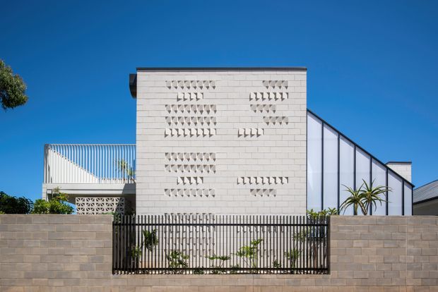 Street-facing exterior of a contemporary multi-level residence behind a low masonry boundary wall and vertical metal gate. The facade combines a pale rendered or concrete-block screen wall with decorative perforated breeze-block detailing, a translucent pitched or angled glazed volume, and a curved metal balcony balustrade. Strong clear daylight and a deep blue sky emphasize the crisp geometric composition.