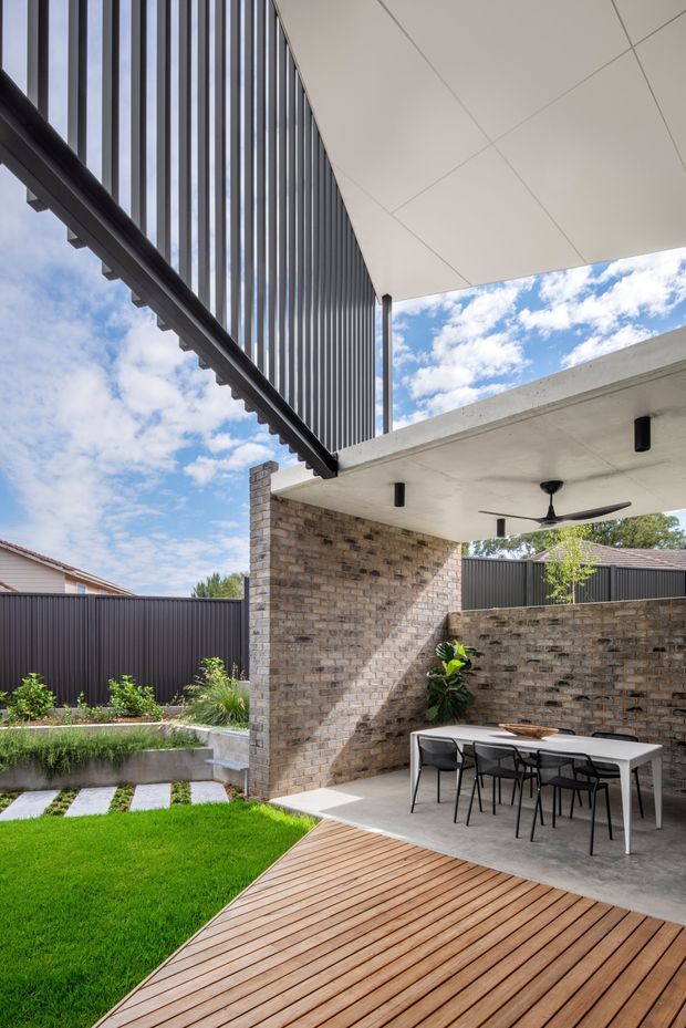 A contemporary covered outdoor dining and entertaining area opening onto a landscaped backyard. The space features a timber deck, polished concrete flooring, exposed light brick walls, dark vertical metal screening, a large white ceiling canopy with recessed downlights and a ceiling fan, and a minimalist outdoor dining table and chairs. Lush lawn and garden beds surround the patio, with bright blue sky visible above.