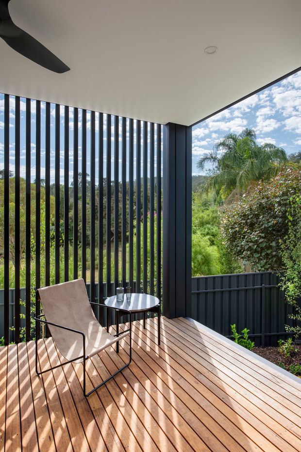 A covered outdoor deck or terrace with warm timber decking, black metal vertical screening and balustrades, and a minimalist black-framed structure. A lounge chair and small round table create a compact seating area, overlooking lush landscaped vegetation and a wooded hillside. Bright natural daylight casts strong linear shadows across the deck.