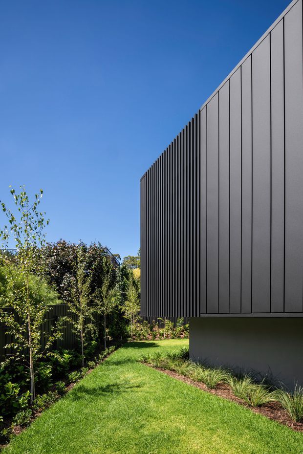 Exterior architectural view of a contemporary building with a sharply projecting upper volume clad in dark grey vertical panels and closely spaced fins. The building sits within a landscaped garden featuring lawn, layered planting and a dark vertical boundary fence, beneath a clear blue sky.