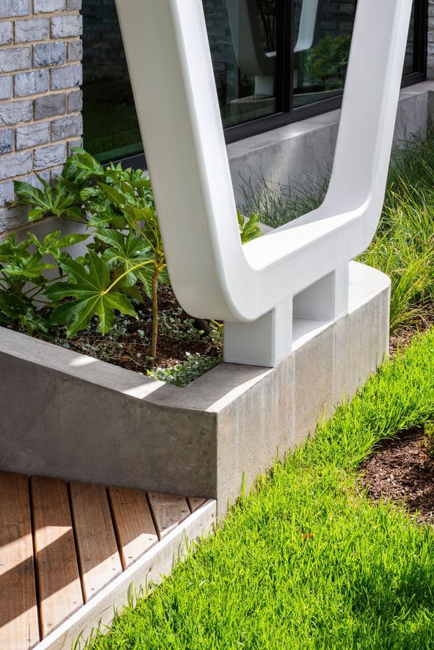 Close-up exterior architectural detail showing a sculptural white painted structural frame or balustrade integrated into a raised concrete garden bed. The setting includes grey brick walls, dark-framed glazing, concrete retaining walls, timber decking steps, and landscaped planting with bright green lawn.