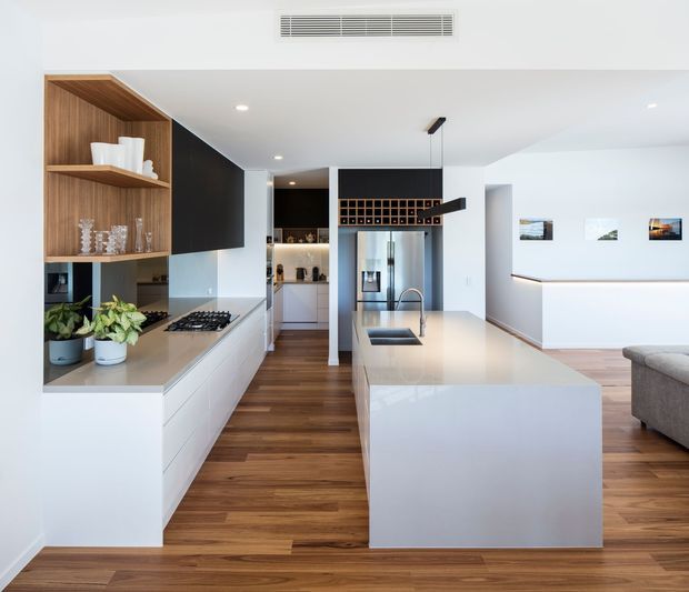 Bright open-plan contemporary kitchen with a large white waterfall island, integrated sink and cooktop, dark cabinetry, timber open shelving, stainless-steel refrigerator, and warm timber flooring. The kitchen opens toward a minimalist living area with white walls and framed artwork.