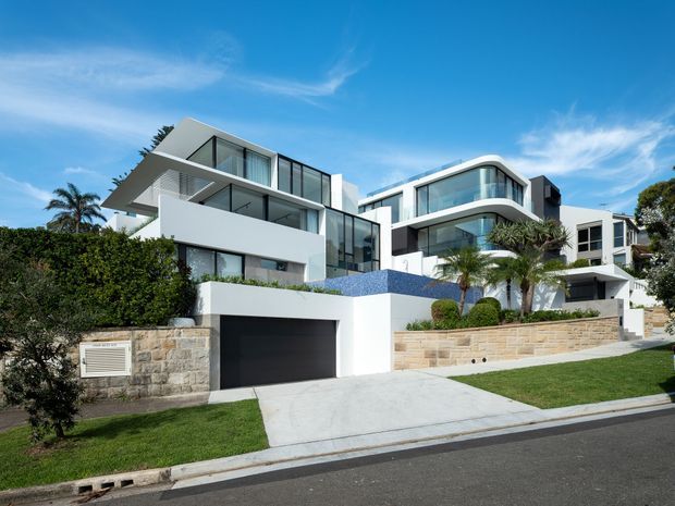 Street-facing view of a large contemporary multi-level residence with crisp white rendered forms, extensive floor-to-ceiling glazing, glass balustrades, projecting balconies, and a curved upper-level corner. The property includes a broad driveway leading to a dark double garage, pale stone retaining walls, manicured planting, and a small blue-tiled pool or water feature visible between the buildings. Bright blue sky and palm planting reinforce the coastal suburban setting.