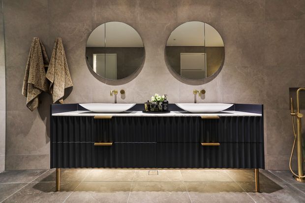 A sophisticated double vanity bathroom with two round mirrors, white vessel basins, a long black fluted vanity with brass handles and legs, and a warm grey stone-look tiled wall and floor. Brass tapware and a freestanding shower fixture add refined contrast, while patterned towels and small greenery provide limited decorative accents.