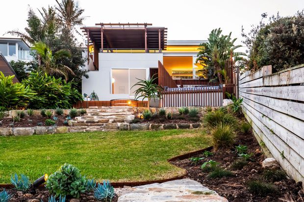 Contemporary two-storey residential facade viewed from a landscaped front garden, featuring white rendered walls, warm timber screening and pergola elements, horizontal yellow-orange accents, a timber front fence, stone garden steps, and lush subtropical planting.