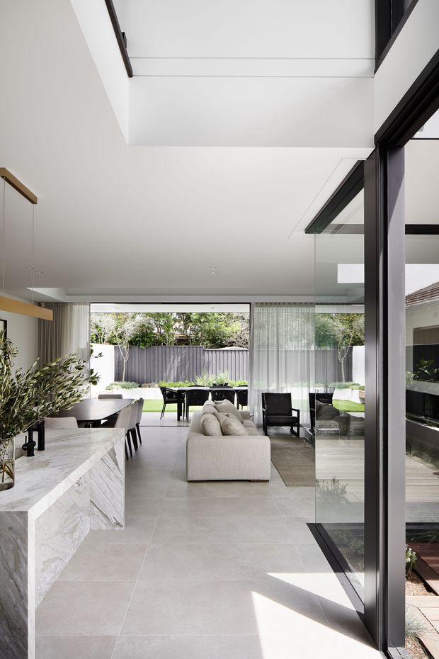 A bright, open-plan contemporary living and dining area connected to a landscaped outdoor courtyard through extensive floor-to-ceiling glazing. The interior features a large cream sectional sofa, dark dining furniture, sheer curtains, pale stone-look tiled flooring, and a prominent white-and-grey veined stone island or built-in bench. Black-framed glass doors and windows create strong indoor–outdoor connection, while a skylight or high-level glazed opening brings additional daylight into the double-height space.