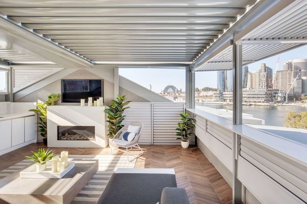 Bright contemporary living room enclosed by a louvred and glazed rooftop terrace structure, with a built-in fireplace and television wall, large windows overlooking a harbour and city skyline, white cabinetry and balustrades, potted plants, and warm herringbone timber flooring.
