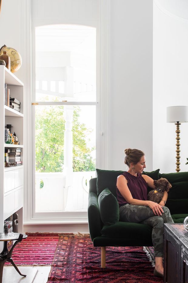 Bright, tall-ceilinged living room with a deep green upholstered sofa, a seated person with a small dog, a patterned red and burgundy rug, white built-in shelving filled with books and objects, a brass-toned floor lamp, and a large white-framed glazed door or window opening to a balcony. The room has an eclectic, richly layered yet airy interior.