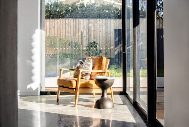 A bright, minimalist interior corner with a mid-century-style upholstered armchair and sculptural side table positioned beside full-height black-framed glazing. Polished grey stone flooring reflects strong sunlight, while a timber privacy fence and planted courtyard are visible outside.