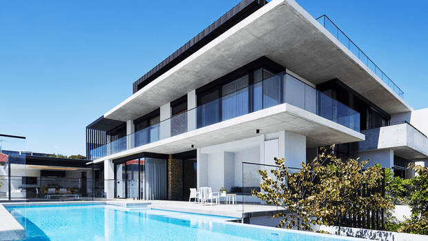 Exterior view of a contemporary multi-level residence with a strong cantilevered concrete slab, extensive dark-framed glazing and glass balustrades, white rendered walls, and a large swimming pool in the foreground. The composition highlights the building facade, outdoor entertaining area, rooftop terrace, and clean geometric lines under a clear blue sky.