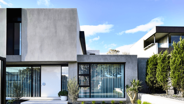 Contemporary multi-level residential facade with smooth grey concrete or rendered walls, expansive dark-framed glazing, a recessed white entry door, and strong rectilinear volumes. The front garden includes sculptural shrubs and drought-tolerant planting, while a neighbouring modern structure is visible at right under a bright blue sky.