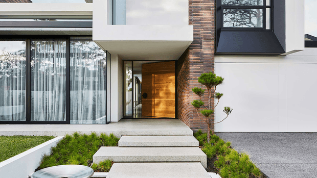 Contemporary residential front entrance with a projecting white geometric canopy, warm timber pivot door, exposed red-brown brick feature wall, extensive black-framed glazing, and crisp white wall panels. Broad concrete stepping slabs lead through a minimalist landscaped garden with sculpted topiary and low planting.