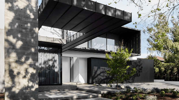 Contemporary multi-level residential exterior with strong geometric composition, dark cantilevered roof and balcony structures, expansive glazing, concrete walls, and a recessed entrance approached by broad steps. A landscaped front garden and mature trees soften the architectural form.