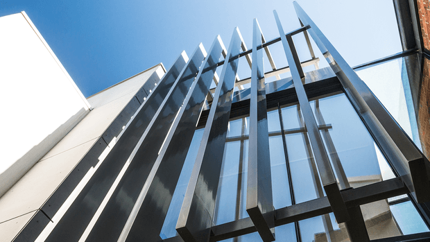 Upward-looking architectural photograph of a narrow contemporary facade or glazed atrium, with tall dark metal fins and exposed structural framing projecting against a clear blue sky. Large vertical glass panels create a dramatic, strongly vertical composition, with adjacent light-coloured and brick masonry walls visible at the edges.