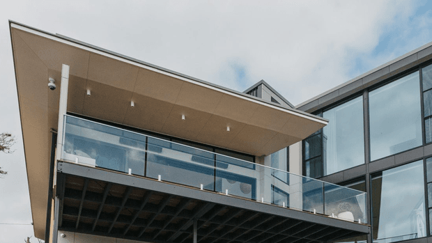 Exterior architectural photograph of a contemporary multi-storey building, viewed from below. The composition features a projecting flat roof canopy with recessed downlights, expansive dark-framed glazing, a glass-balustraded balcony, and a dark exposed structural soffit beneath the cantilevered terrace. A small pitched roof element is visible behind the main flat-roofed volume.