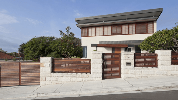 Street-facing exterior of a contemporary two-storey residence with a broad low-pitched roof, cream rendered upper walls, white masonry boundary walls, and extensive warm timber slat screening across the garage, pedestrian gate, balcony, and front fence. The property has a paved driveway and footpath, landscaped shrubs, and a clean urban suburban setting.