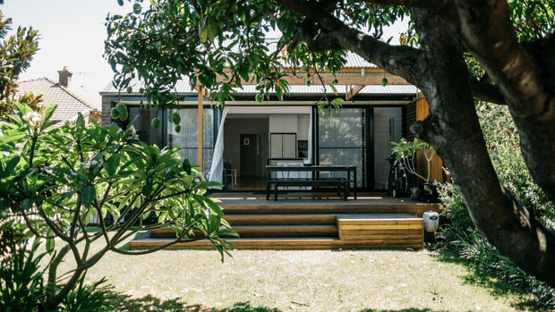 A compact modern single-storey backyard dwelling or extension viewed from a landscaped garden. The building has a low-pitched corrugated roof, broad glazed openings with dark frames, timber screening and cladding, and a raised timber deck with several steps. A dark outdoor dining table sits beneath the covered terrace, while mature trees and tropical planting frame the view. A larger tiled-roof house is partially visible behind it.