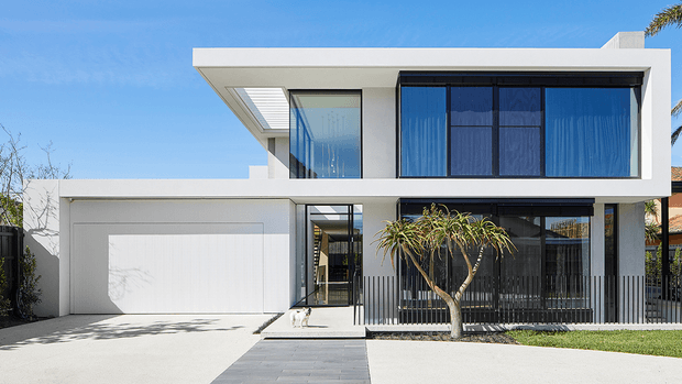 Street-facing view of a contemporary two-storey residence with a crisp white rendered facade, strong horizontal roof and balcony lines, extensive dark-framed glazing, a recessed upper-level terrace, and a minimal front garden. A broad white garage door and paved driveway sit to the left, while a small sculptural tree and black vertical metal fencing define the entry frontage. The composition is clean, rectilinear, and brightly lit under a clear blue sky.